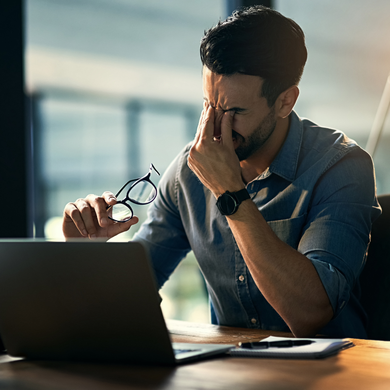 An employee rubs their eyes in stress at their work desk.